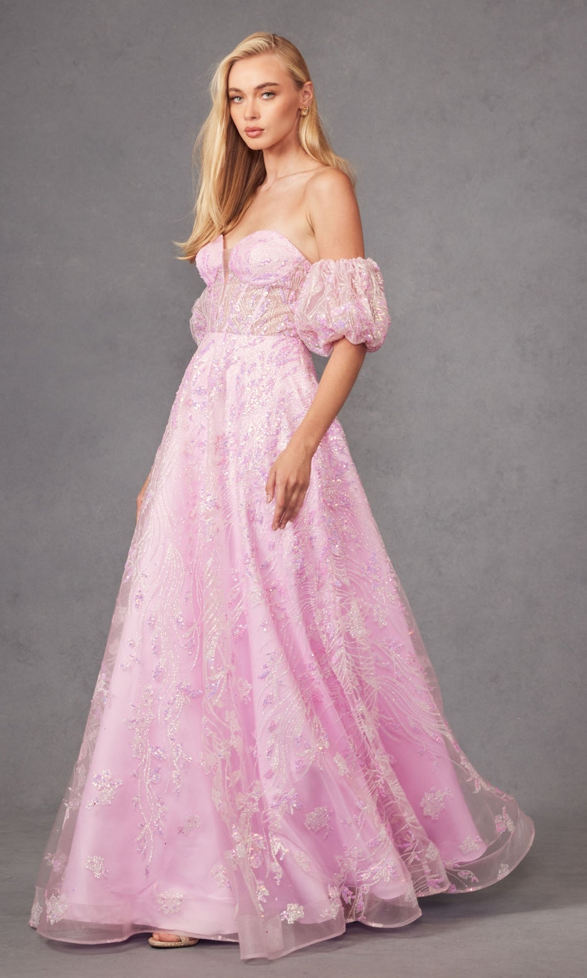 A woman stands against a gray background wearing the Puff-Sleeve Strapless Long Glitter Prom Dress 2404, featuring light pink fabric, sheer puff sleeves, and sparkling embroidery.